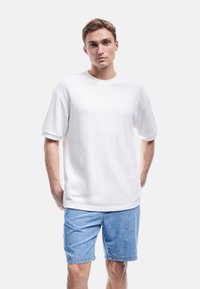 Weißes T-Shirt mit kurzen Ärmeln, runder Ausschnitt und lockerer Passform, kombiniert mit hellblauen Denimshorts, was einen lässigen und bequemen Stil zeigt.