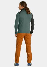 Grüner und schwarzer Reißverschluss-Pullover mit langen Ärmeln, kombiniert mit orangenen Hosen. Aus strukturiertem Stoff mit enganliegendem Design. Die Schuhe sind grau und blau.