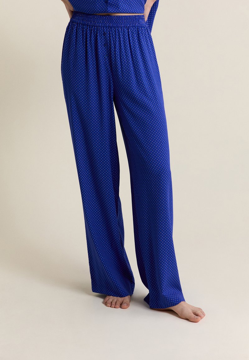 Mujer con pantalones de pijama azules y sueltos con un pequeño patrón de puntos, de pie descalza contra un fondo liso.