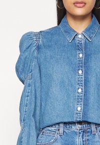 Blue denim shirt with a button-up front, collar, and puffed sleeves. Soft texture and subtle fading with small buttons and a slight shine.