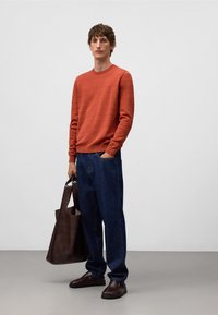 Uomo che indossa un maglione arancione ruggine, jeans blu scuro, scarpe di pelle marrone e tiene una grande borsa a tracolla in pelle marrone, in piedi contro un muro bianco.