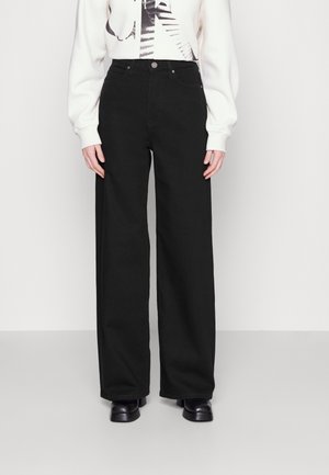 Persona con jeans negros de tiro alto y pernera ancha, camiseta blanca de manga larga con gráfico negro y zapatos negros, de pie frente a un fondo liso.