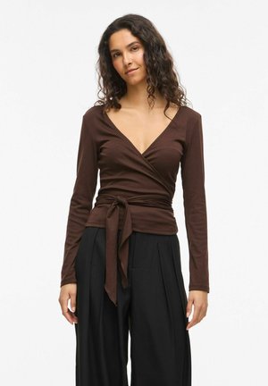 Žena s dlhými kučeravými vlasmi, ktorá má na sebe hnedé dlhé rukávy, zviazané v páse, a voľné čierne nohavice, stojaca na bielom pozadí.