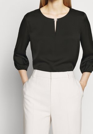 Sort silk bluse med rund halsudskæring, center nøglehulskæringsdetalje og sølv cirkulær accent. Har trekvart puffærmer. Parret med hvide bukser.