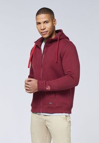 Bordeauxfarbener Zip-Hoodie mit roten Kordeln, ausgestattet mit seitlichen Taschen und einem Logodetail am Bündchen, aus einem strukturierten Stoff gefertigt.