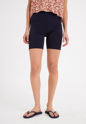 Femme portant un short bleu marine cintré, un haut rouge à motif floral et des tongs bleu marine, debout devant un fond blanc uni.