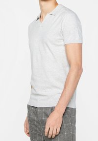 Camiseta polo gris de manga corta con cuello en V, hecha de una tela suave. Presenta una silueta ajustada y puños acanalados. Combinada con pantalones grises de cuadros.