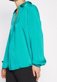 Teal silk blouse with a narrow collar, long cuffed sleeves, and a relaxed fit. The fabric has a smooth, glossy texture.