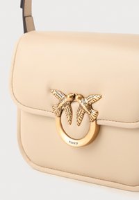 Bolso de hombro de cuero beige con cierre de metal dorado que presenta dos pájaros detallados frente a frente sosteniendo un anillo circular.