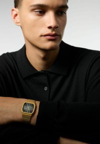 Montre bracelet en or avec un boîtier rectangulaire et un cadran noir. Le bracelet présente un design texturé et maillé. Le modèle porte un polo noir.