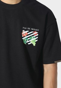 Czarna bawełniana koszulka z grafiką na klatce piersiowej przedstawiającą czerwone róże, zielone winogrona oraz paski. Tekst "PAS DE MONACO" nad wzorem.