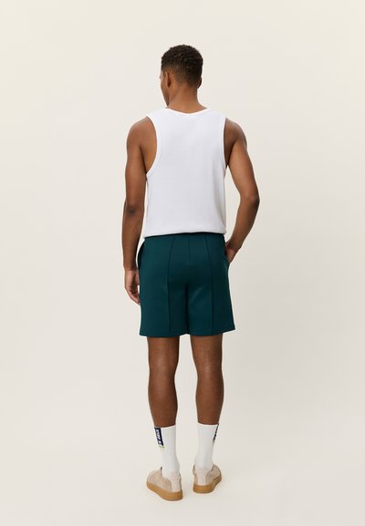 Débardeur blanc sans manches associé à un short vert foncé avec poches latérales. Porté avec des baskets couleur crème et des chaussettes blanches.