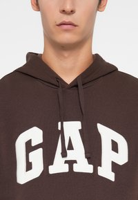 Brown hoodie with white "GAP" lettering on the front, featuring a pullover design and drawstrings at the neckline. Soft texture.