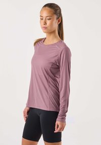 Pitkähihainen urheilupaita hillityssä luumunvärissä, jossa on teksturoitu kangas ja heijastavia yksityiskohtia, yhdistetty mustiin, vartalonmyötäisiin shortseihin.