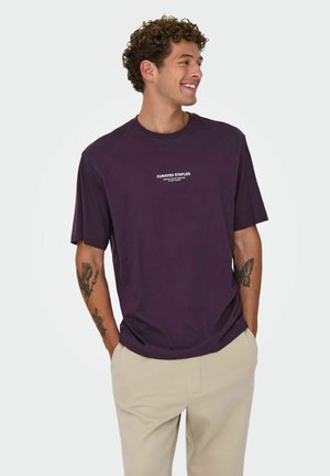 Lilla kortærmet t-shirt med teksten "CURATED STAPLES" i hvid på forsiden. Fremstillet af bomuld, afslappet pasform, parret med lyse beige bukser.