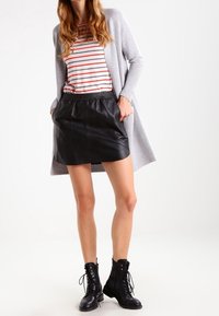 Vrouw in een grijze lange cardigan, een rood-wit gestreept shirt, een zwarte leren rok en zwarte veterschoenen, staand met de handen in de zakken.