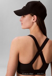 Femeie purtând un sutien sport negru Calvin Klein cu bretele încrucișate pe spate și o șapcă neagră, văzută din spate pe un fundal gri.