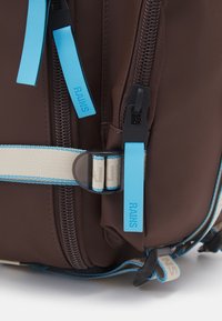Brauner Rucksack mit blauen Akzenten, aus strukturiertem Material, mit Reißverschlüssen, verstellbaren Trägern und einem auffälligen Markenschild.