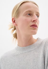 Pulover tricotat în gri deschis, cu un guler rotund ribatat. Textură fină și ajustare lejeră. Modelul are o coadă de cal joasă și un machiaj neutru.