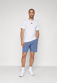 Bela športna majica z črnim logotipom, v kombinaciji z modrimi kratkimi hlačami. Model stoji v belih športnih čevljih, kar prikazuje sproščen fit in sproščen stil.