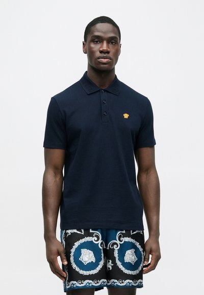 Μπλε σκουρό πόλο με γιακά και τρία κουμπιά, με ένα μικρό χρυσό έμβλημα. Συνδυασμένο με σορτς σε ασπρόμαυρο και μπλε μοτίβο.