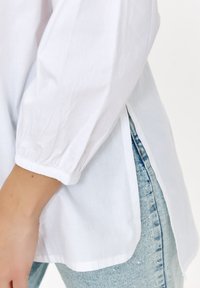 Biała koszula z długim rękawem, z marszczonymi mankietami i bocznymi rozcięciami, w zestawieniu z jasnoniebieskimi spodniami dżinsowymi z delikatnymi zdobieniami.