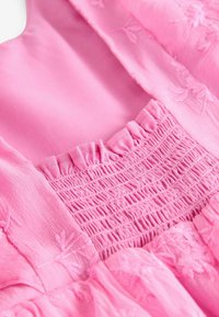 Roze jurk detail met een gesmokt lijfje, ruches bij de halslijn en geborduurde bloemenpatronen op een gestructureerde, lichtgewicht stof.