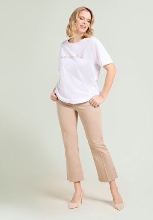 Donna bionda in piedi con le mani nelle tasche che indossa una camicia bianca a maniche corte con testo, pantaloni beige corti e tacchi alti beige su sfondo chiaro.