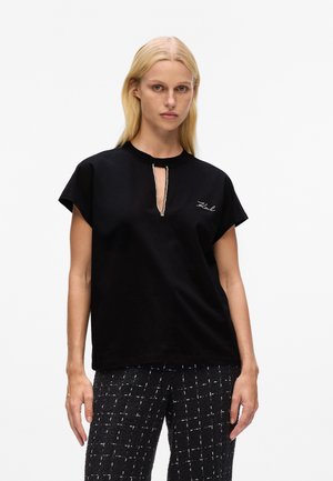 Blonde woman wearing a black short-sleeve top with a narrow front slit and embroidered text, paired with black and white checkered pants.