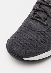 Närmre bild av tån på en svart stickad sneakersko med textilmaterial, sydd möbel av mocka för snörpanelen, svarta snören och vit gummi sula.