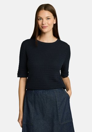 Junge Frau mit glatten braunen Haaren trägt ein strukturiertes schwarzes Kurzarmoberteil und einen dunklen Jeansrock mit den Händen in den Taschen.