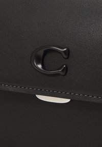 Bolso de cuero negro con un brillante logo "C" en negro, con un acento blanco contrastante y costuras sutiles a lo largo del borde.