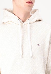 Cream hoodie with geometric pattern, white drawstrings, silver hardware, and a small logo accent on the chest. Soft texture.