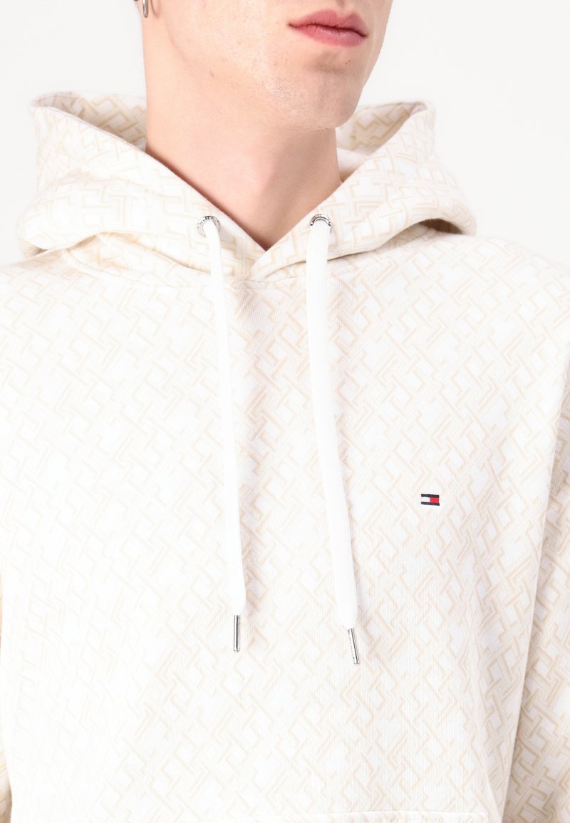 Cremfarbener Hoodie mit geometrischem Muster, weißen Kordeln, silbernen Beschlägen und einem kleinen Logo-Detail auf der Brust. Weicher Stoff.