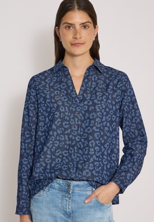 Femme aux longs cheveux foncés porte un chemisier bleu à manches longues à motifs et un jean bleu clair, la main droite dans la poche.
