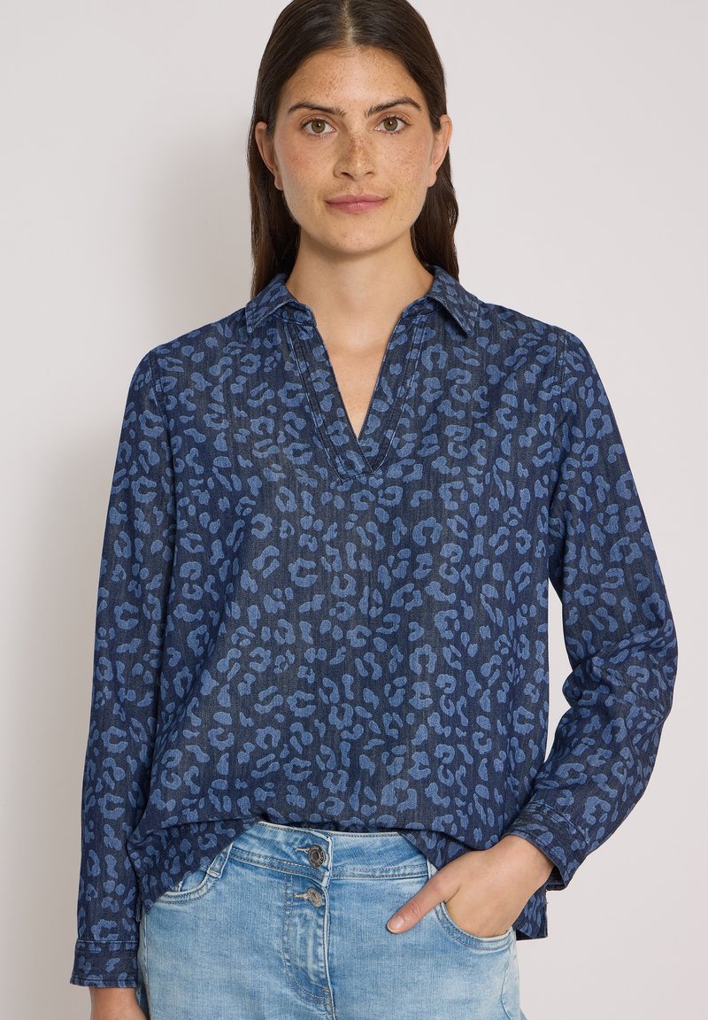 Femme aux longs cheveux foncés porte un chemisier bleu à manches longues à motifs et un jean bleu clair, la main droite dans la poche.