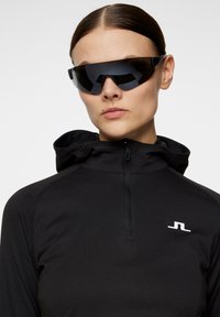 Svart zip-up hoodie med hög krage och vitt logo på vänster bröst. Modellen bär stora svarta solglasögon med minimalistisk design.