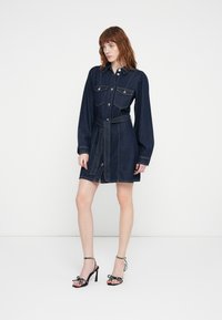 Robe chemise en denim foncé avec deux poches poitrine, fermeture à boutons et ceinture à nouer. Associée à des talons noirs avec des accents à pois.