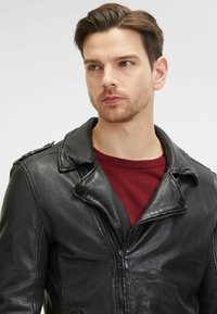 Giacca di pelle nera con colletto a scollo, chiusura con zip e spalline. Indossata sopra una camicia bordeaux. Tessuto liscio con un design aderente.