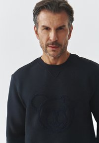 Sudadera azul marino de tela suave con un logo de oso texturizado en la parte delantera. Cuello redondo y puños acanalados.