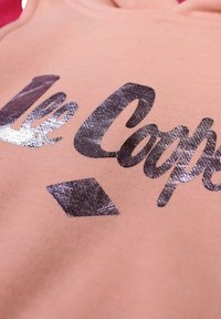 Tissu pêche clair à texture lisse, orné d'un texte métallique argenté affichant "Le Coops" et d'un accent en forme de losange en dessous.