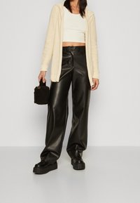 Žena ve krémovém pleteném svetru, bílém crop topu, černých širokých kožených kalhotách, na platformě, držící malou černou texturovanou kabelku.