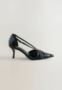 Black patent leather high heel shoe with a pointed toe, crisscross straps, and a circular gold accent on the strap. Smooth texture.