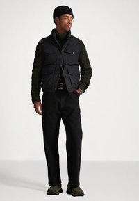 Juodas liemenė su aukštu apykakliu ir keliais priekiniais kišenėmis, dėvimas ant tamsiai žalios pintos megztinio, derinamas su juodomis kelnėmis ir alyvuogių spalvos sportbačiais.