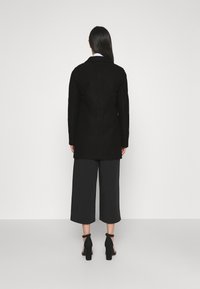 Donna con capelli lunghi e lisci che indossa un cappotto nero lungo fino al ginocchio, pantaloni a gamba larga accorciati e sandali neri con tacco alto, vista da dietro.