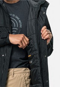 Zwarte pufferjack met een ritssluiting aan de voorkant en drukknopen, met een gestikte binnenkant en logo-patch, gedragen over een donker T-shirt en beige broek.