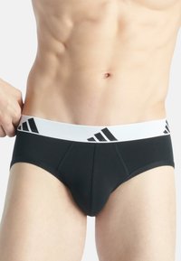 Schwarze Baumwollslips mit einem weißen elastischen Bund, der mit schwarzem Adidas-Branding versehen ist. Das Design ist tailliert und hat eine glatte Textur und Nähte.