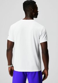 White short-sleeve t-shirt with a round neckline, smooth texture, and logo detail on the left sleeve. The model is wearing purple shorts.