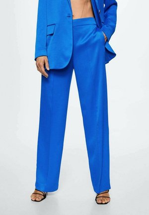 Person wearing bright blue wide-leg trousers with matching blazer and black strappy high-heeled sandals, standing with one hand in pocket.