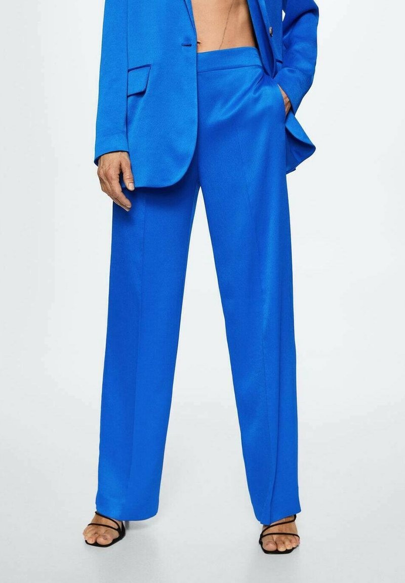 Personne portant un pantalon large bleu vif avec un blazer assorti et des sandales à talons hauts noires à brides, debout avec une main dans la poche.